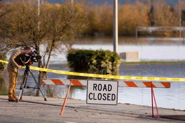 A person films a flooded street behind a "Road Closed" sign and caution tape; trees and water are visible in the background.