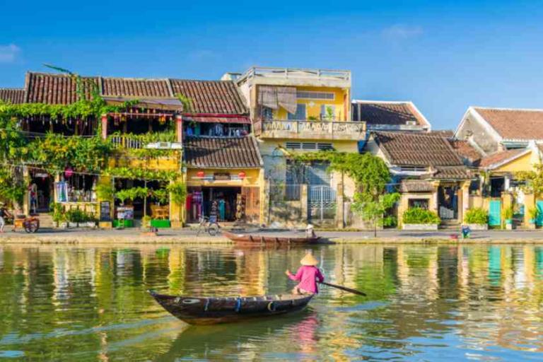 A person wearing a conical hat rows a small wooden boat on a calm river, with colorful old buildings and greenery lining the riverside under a clear blue sky.