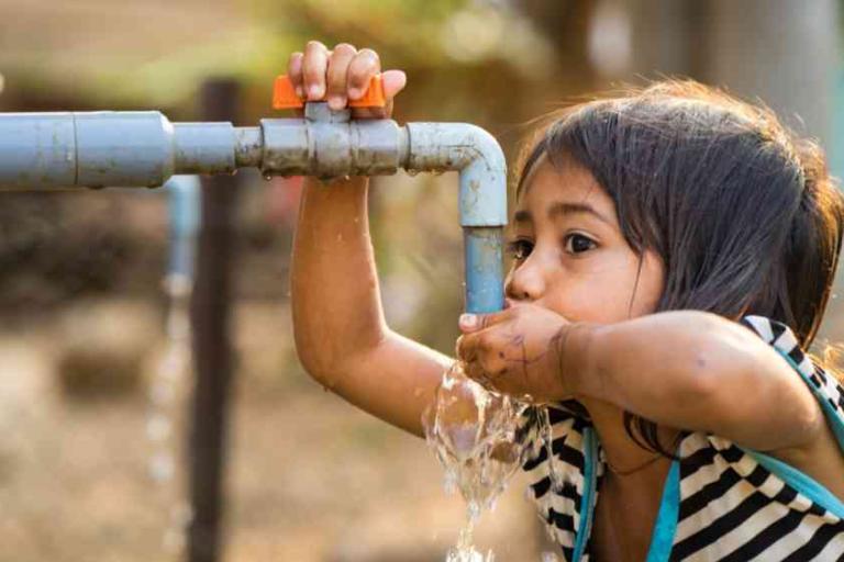 A young girl drinks water from a blue outdoor faucet, using one hand to hold the tap and the other to catch the water.