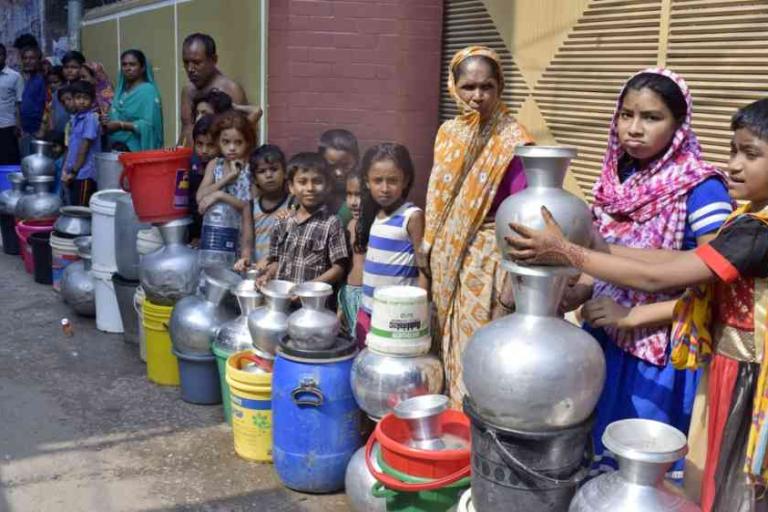 A group of people, including women and children, stand in line with metal and plastic containers, waiting to collect water on a street.