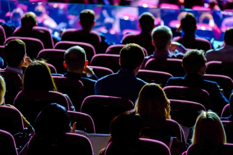 Audience members are seated in rows of theater-style chairs, facing a stage or screen, with dim lighting in the room.