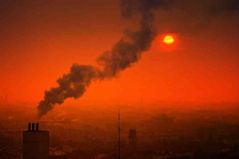 Industrial chimney emitting dark smoke into an orange sky at sunset, with the sun partially obscured by haze over a cityscape.