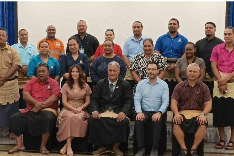 A group of men and women pose together indoors, with most men wearing traditional Tongan attire and some people in business or casual clothes, seated and standing in rows.