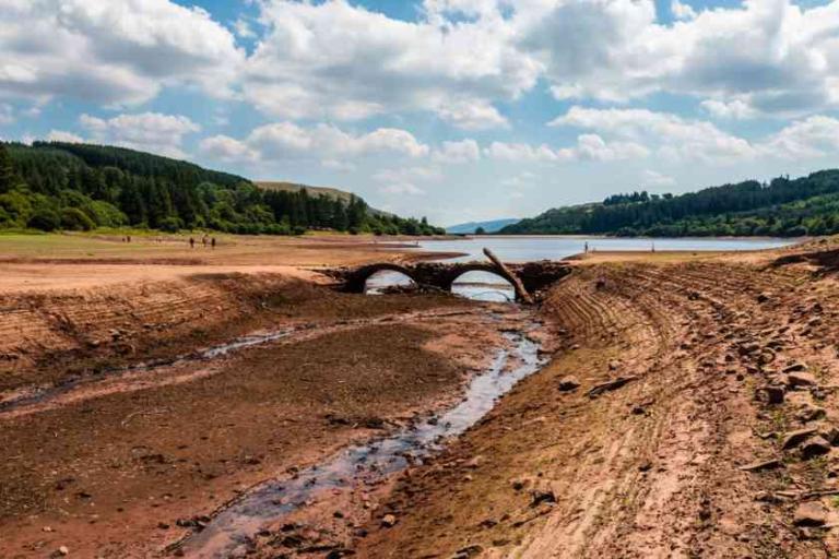 A dried-out riverbed reveals an old stone bridge, surrounded by exposed earth, with a lake and forested hills in the background under a partly cloudy sky.