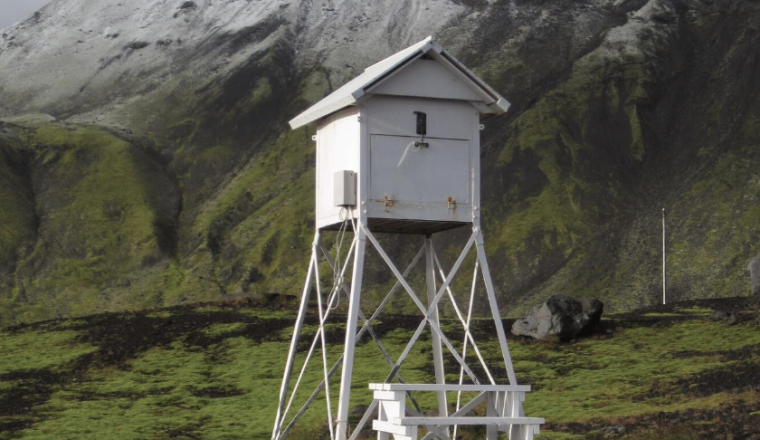 A white watchtower sits in the middle of a field with mountains in the background.