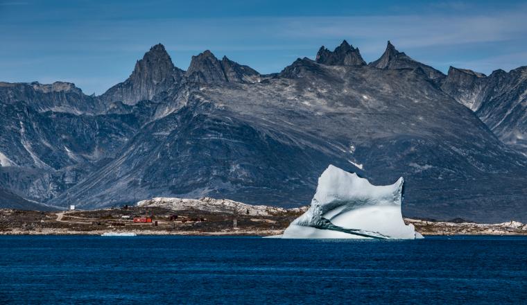 A large iceberg floats in a calm blue sea with rocky mountains in the background under a clear sky.