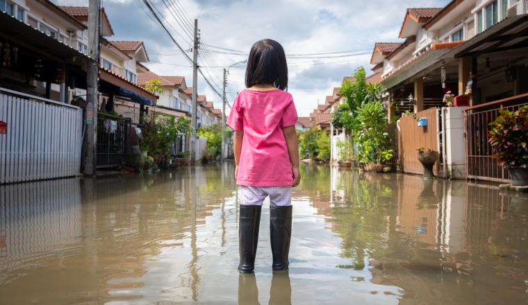 A child in a pink shirt and black boots stands in the middle of a flooded residential street surrounded by houses on a cloudy day.