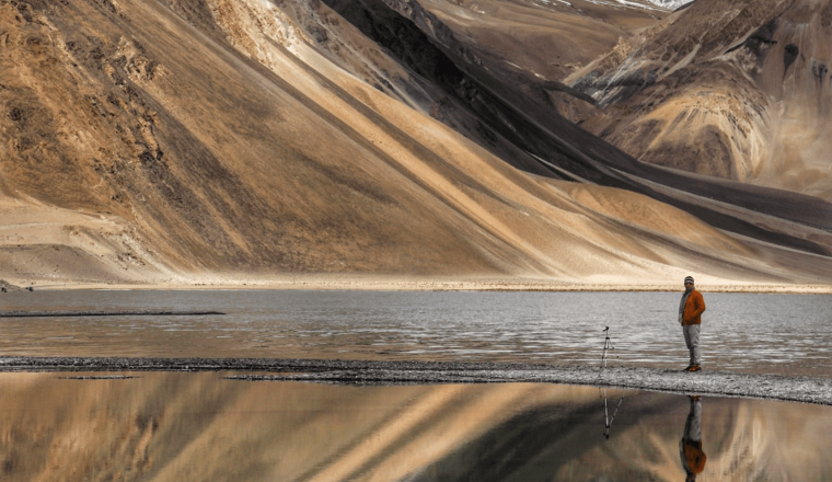 A person stands near a tripod by a reflective mountain lake with snow-covered peaks in the background.