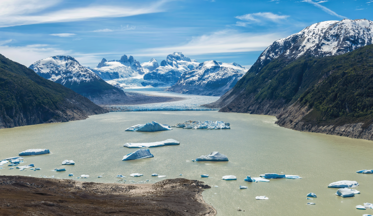 A glacier-fed lake with floating icebergs, bordered by rugged mountains and snow-capped peaks under a partly cloudy sky.
