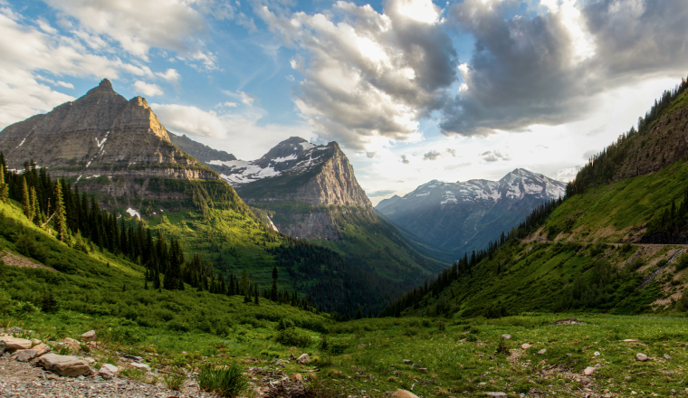 Mountain valley view with grassy slopes, coniferous trees, and snow-capped peaks under a partly cloudy sky. A narrow road is visible on the right hillside.