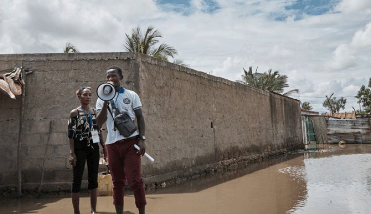A person stands in floodwater using a megaphone, while another individual stands nearby. A partially flooded building is in the background under a cloudy sky.