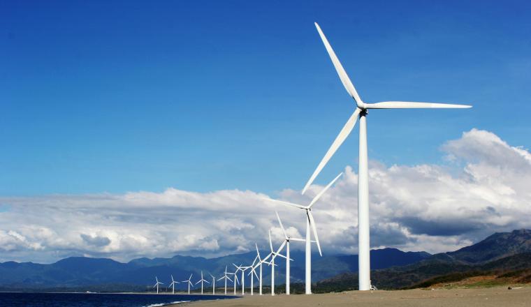 A row of wind turbines stands along a sandy coastline with mountains and blue sky in the background.