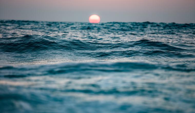 The sun sets over a calm ocean, with gentle waves in the foreground and a pinkish sky on the horizon.