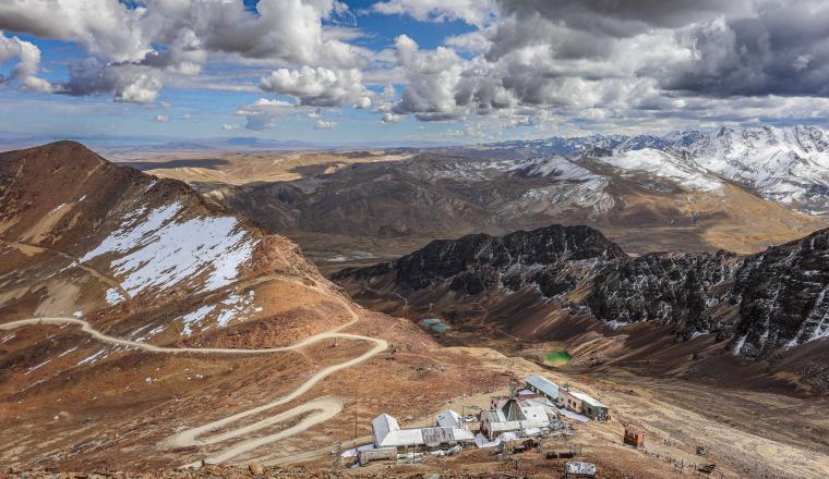 Mountain landscape with a winding road leading to buildings at the base, surrounded by rocky hills, snow patches, and distant snow-capped peaks under a cloudy sky.