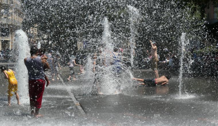 People, including children, play and cool off among multiple water fountains in an urban outdoor setting on a sunny day.