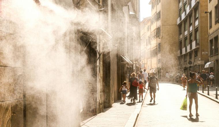 People walk down a sunlit street as mist or steam sprays from a building on the left, creating a hazy effect in the air.