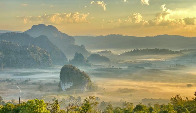 A scenic landscape with mountains, rolling hills, and mist covering the valley under a partly cloudy sky at sunrise.