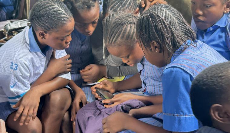 A group of schoolgirls in uniforms sit closely together, looking intently at a smartphone, inside a classroom.