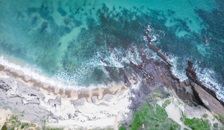 Aerial view of a rocky shoreline with waves crashing onto a sandy beach and clear turquoise water.