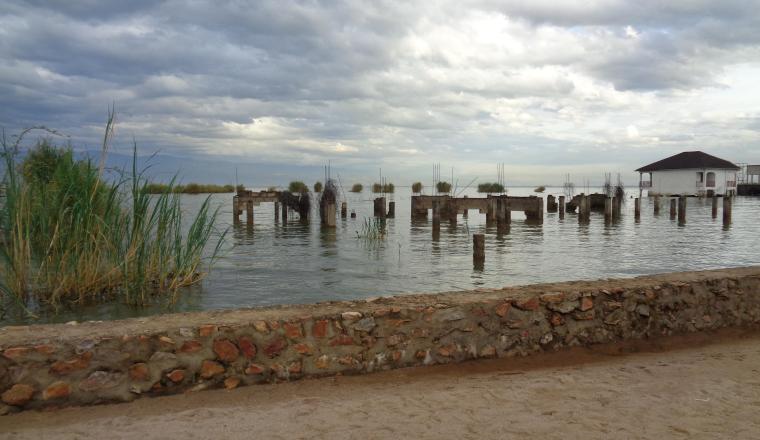 Sandy path with a stone wall, water with old wooden pilings, reeds on the left, and a small white building on the right under a cloudy sky.