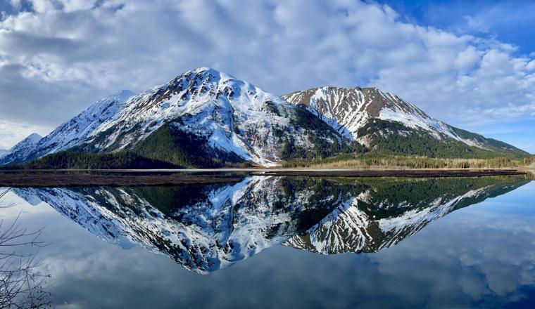 Snow-capped mountains are reflected in a calm lake under a blue sky with scattered clouds.