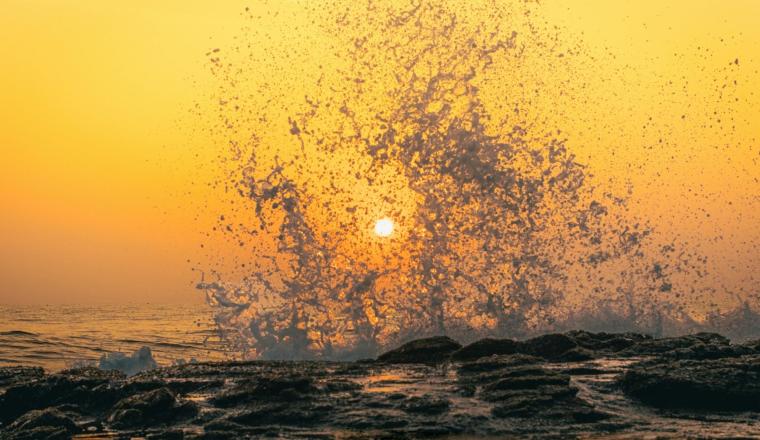 Ocean waves splash against rocks at sunset, with the sun visible through water droplets and an orange sky in the background.