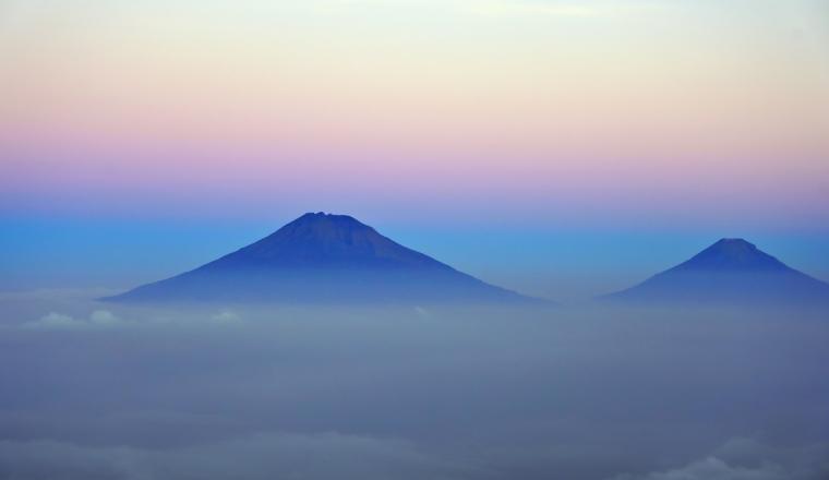 Two mountain peaks rise above a layer of clouds at sunrise, with a gradient sky transitioning from pink to blue in the background.
