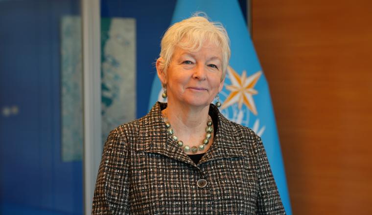A woman with short, white hair wearing a patterned blazer and a beaded necklace, standing in front of a blue flag.