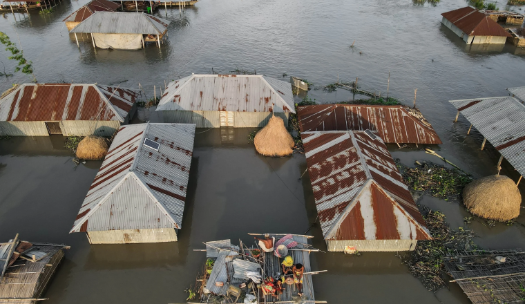 Aerial view of houses partially submerged in floodwater, with people on a makeshift raft in the center.