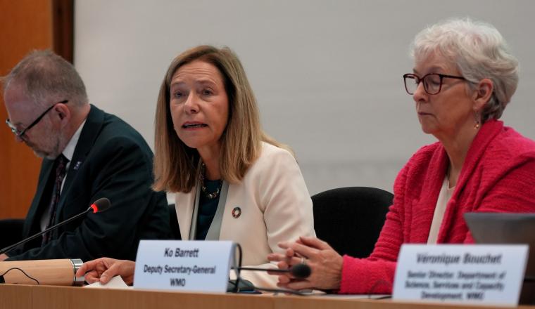 Three people seated at a table with nameplates during a meeting; Ko Barrett sits in the center.