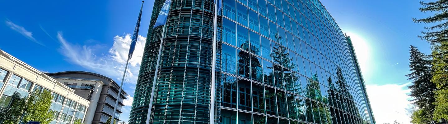 A modern glass office building with a curved facade stands under a clear blue sky, surrounded by trees and neighboring structures.