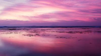 A pink and purple sky is reflected in the water on the beach.