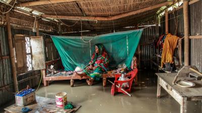 A person sits on a bed under a mosquito net inside a flooded wooden and metal hut. Household items are scattered, and water covers the floor.