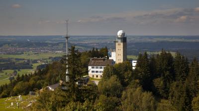 Aerial view of a hilly landscape with a meteorological tower, two buildings, forested areas, and distant fields on a clear day.