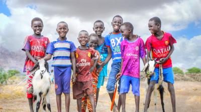 A group of seven children stands outdoors on a dirt path, smiling at the camera. Two of the children are holding goats. The sky is partly cloudy in the background.