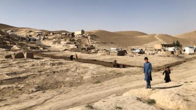 A man and a child walk on a dirt path in a rural, arid landscape with simple housing structures and people working in the distance.