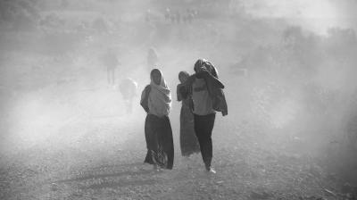 A group of people walk on a dusty path in an arid landscape, with some covering their heads and faces. The visibility is low due to the dust in the air.