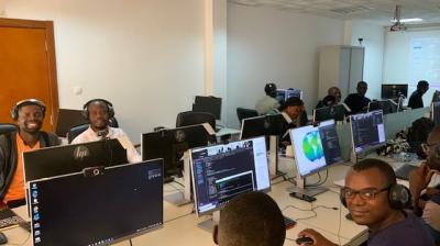 A room with multiple people wearing headphones and working on computers. Some screens show coding or data visualization programs. The environment appears to be collaborative and focused.