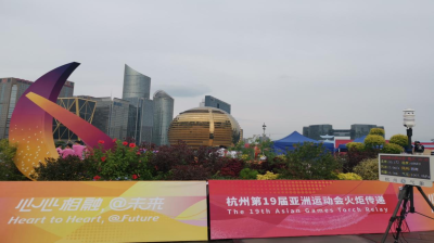 The image shows a public display with banners and signage for the 19th Asian Games Torch Relay in Hangzhou, including the slogan "Heart to Heart, @ Future," set against urban buildings and greenery.