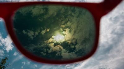 View of cloudy sky with a bright sun seen through red-framed sunglasses.