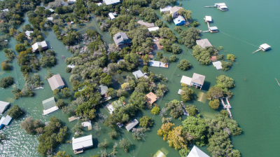Aerial view of a flooded community with partially submerged houses and trees, surrounded by rising water levels. Some structures are completely surrounded by water.