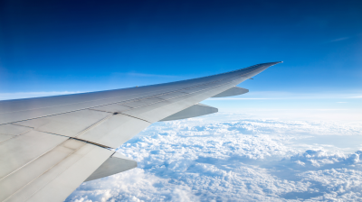 View of an airplane wing flying over a layer of clouds with a clear blue sky in the background.