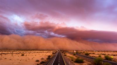 A long road stretches into the horizon under a dramatic sky filled with an approaching dust storm at sunset.