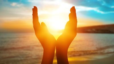 Hands are cupped together in front of a sunset over a beach, framing the sun. The sky has vibrant shades of orange, yellow, and blue.