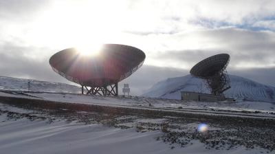 Two large satellite dishes are situated in a snowy landscape with mountains in the background. The sun is partially visible behind one of the dishes, casting light through its structure.