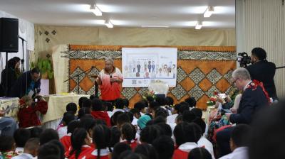 A speaker addresses a group of schoolchildren seated on the floor in a decorated room with a projected presentation behind them. Several adults are standing, and a cameraman is filming the event.