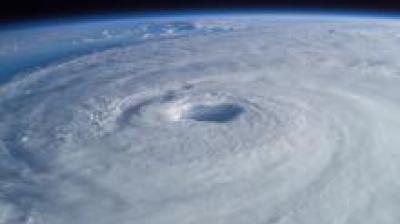 A satellite image of a large, spiraling hurricane with a clearly visible eye at the center, taken from space, showing the swirling cloud patterns over the Earth.