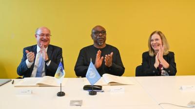 Three people sit at a table with documents and small flags in front of them, clapping their hands with smiles. The background is a yellow wall.