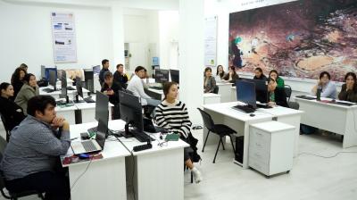 A classroom with several students sitting at desks with computers. They are attentively facing the front where an instructor is visible. A large, detailed map is displayed on the wall.