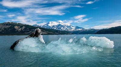 A partially submerged iceberg floats in a tranquil blue lake with snow-capped mountains and a clear sky in the background.